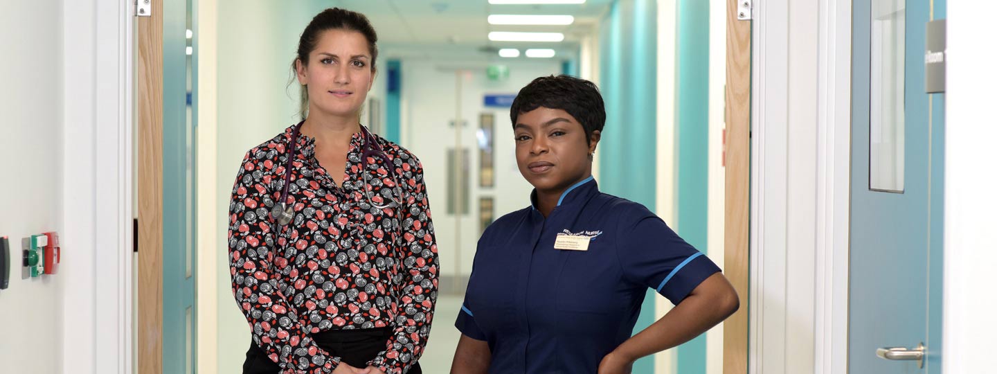 Two healthcare professionals looking at the camera stood in a hospital corridor.