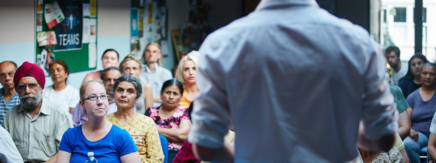 A man standing at the front of the room who is talking to a group of people.