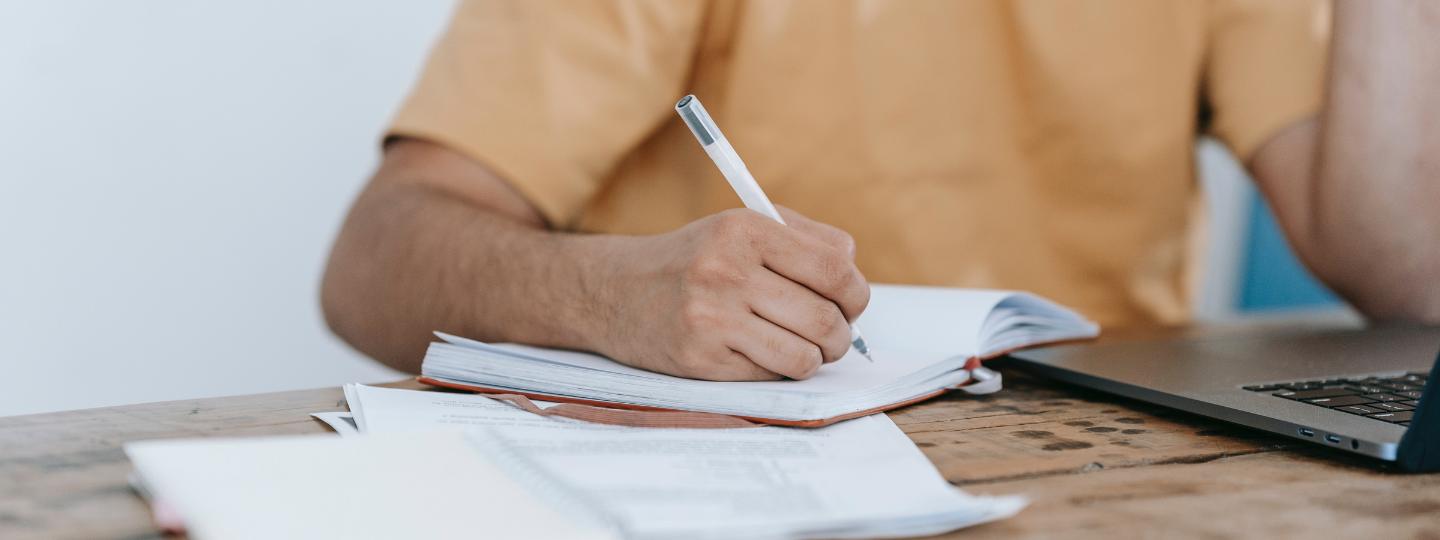 Man writing in book beside laptop