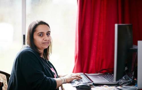 Priti working on her computer in her living room beside a red curtain