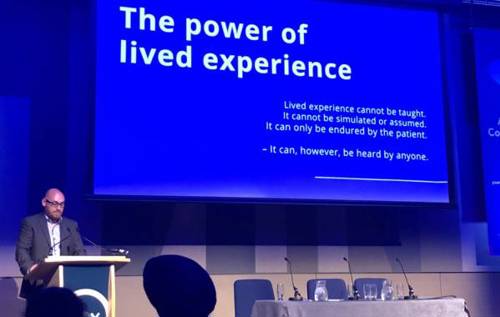 Man speaking at conference in front of Powerpoint slide which reads 'The power of lived experience'