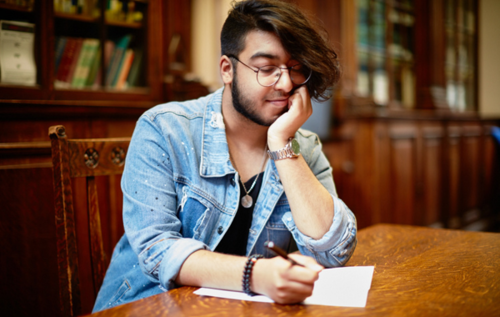 Adam thinking and writing in library with lots of wooden furniture and books