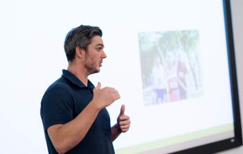 Male researcher wearing black polo t-shirt speaking empathically about ASCOT trial in front of slideshow