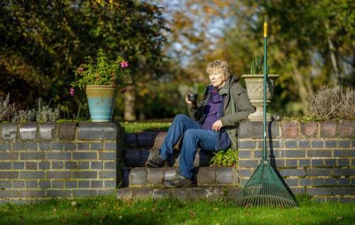 Blonde woman wearing outdoor clothes sat on step drinking a cup of tea in her garden beside a rake