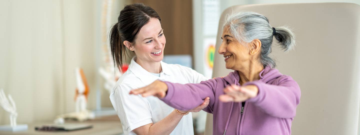 Smiling nurse helping woman wearing pink jumper to raise arms 