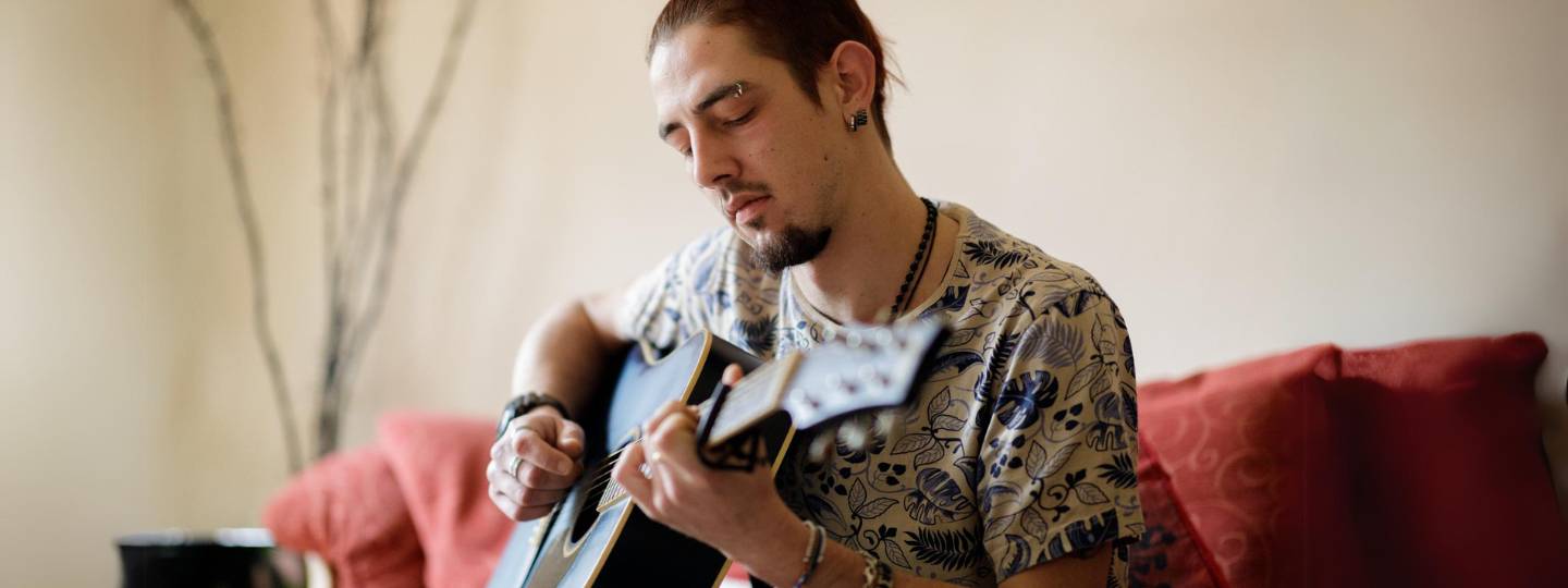 Man with piercings playing guitar on red sofa