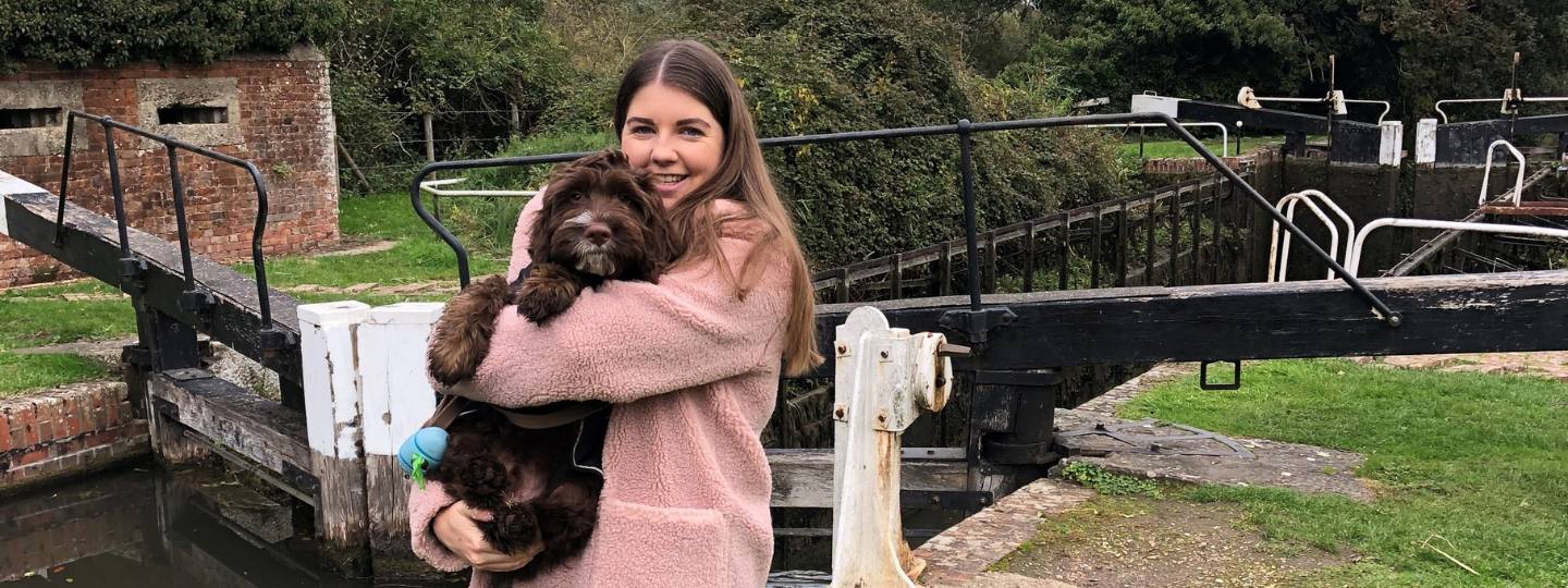 Smiling Lauren wearing pink fluffy coat holding a brown spaniel in front of a navigation lock