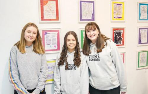 Three young people standing in front of artwork wearing Versus Arthritis jumpers