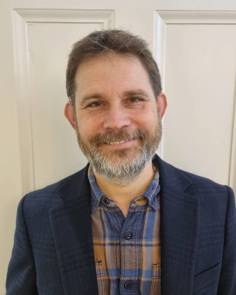 Smiling photo of Jonathan who has a beard and is wearing plaid shirt and suit jacket