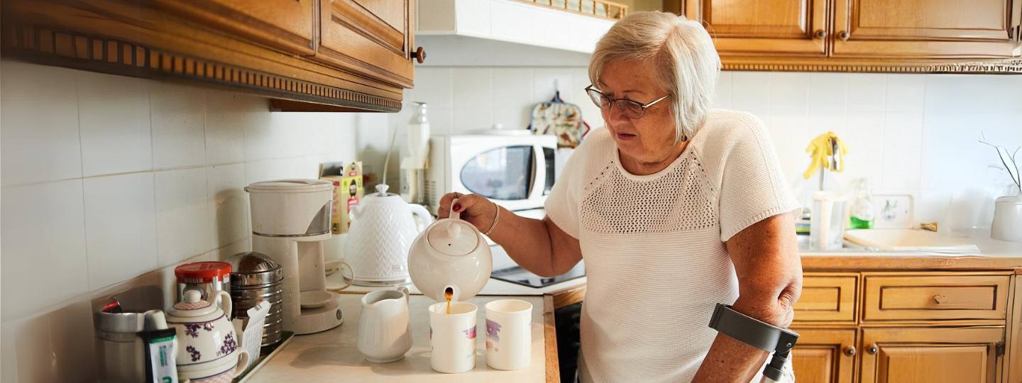 Christine wearing white top and using crutch while making two cups of tea in her kitchen