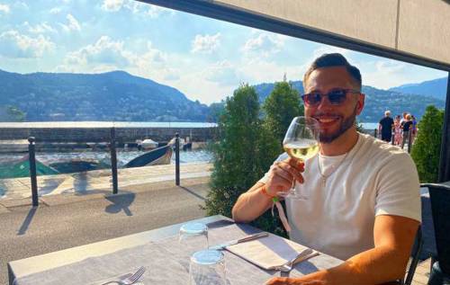 Smiling James wearing sunglasses and drinking a glass of white wine in a sunny restaurant outdoors with mountaintops in the background