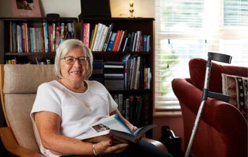 Smiling Christine sitting down in living room reading a book beside her crutch